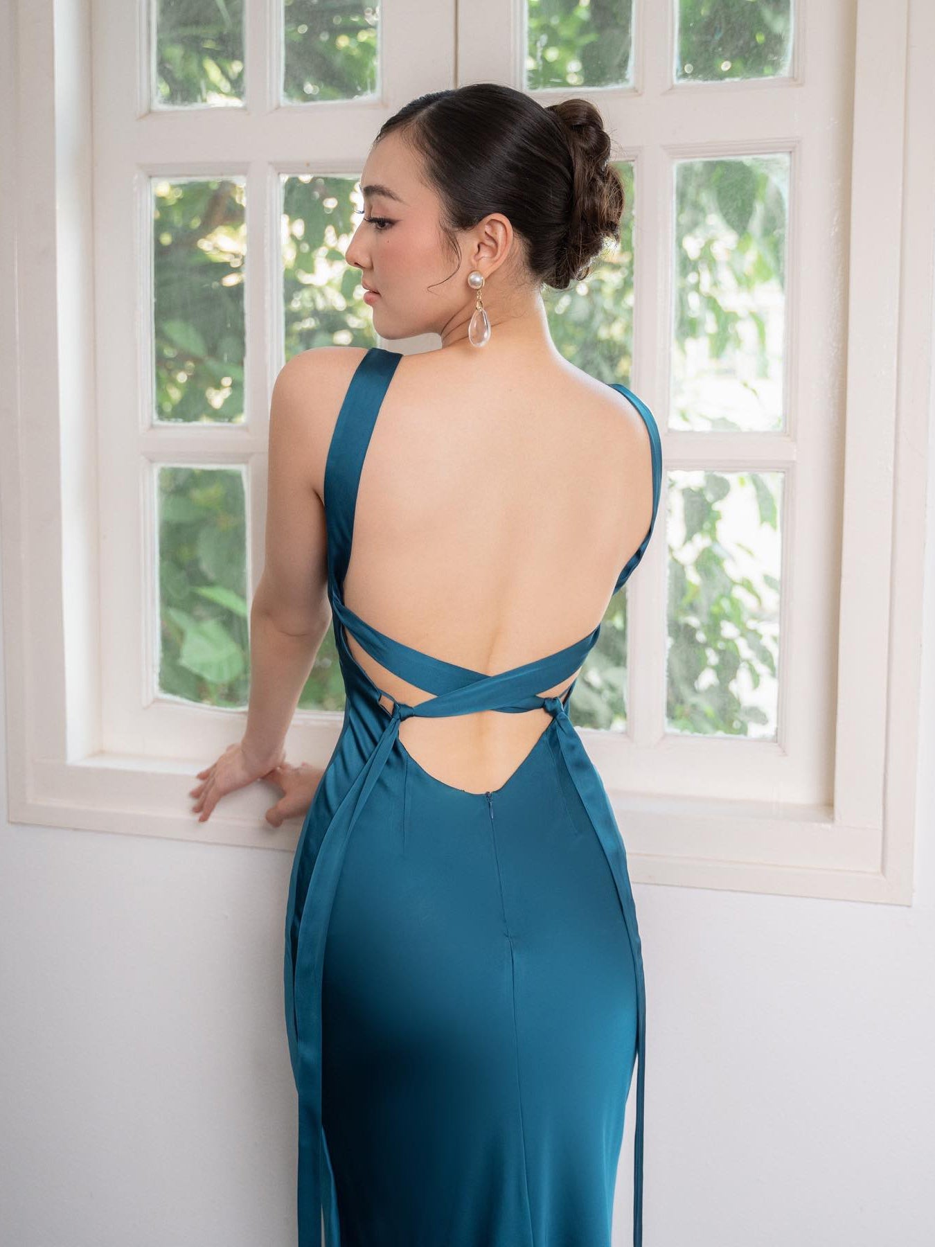 Donna in abito da sera blu con schiena scoperta, davanti a una finestra. Eleganza, moda femminile, abito lungo, stile sofisticato.