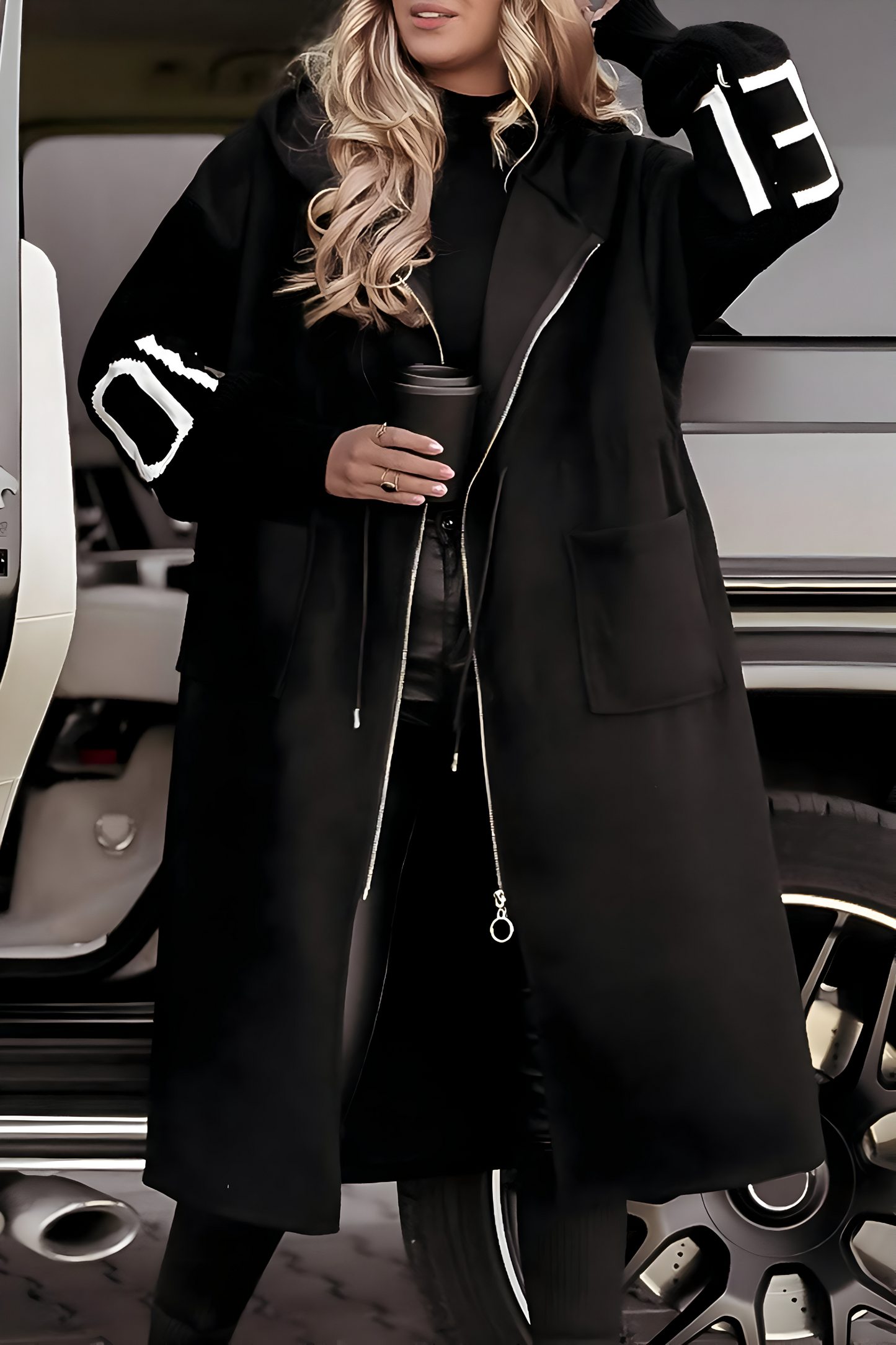 Donna con cappotto nero lungo, dettagli bianchi sulle maniche, tiene una tazza di caffè. Moda invernale, look casual chic, auto di lusso sullo sfondo.