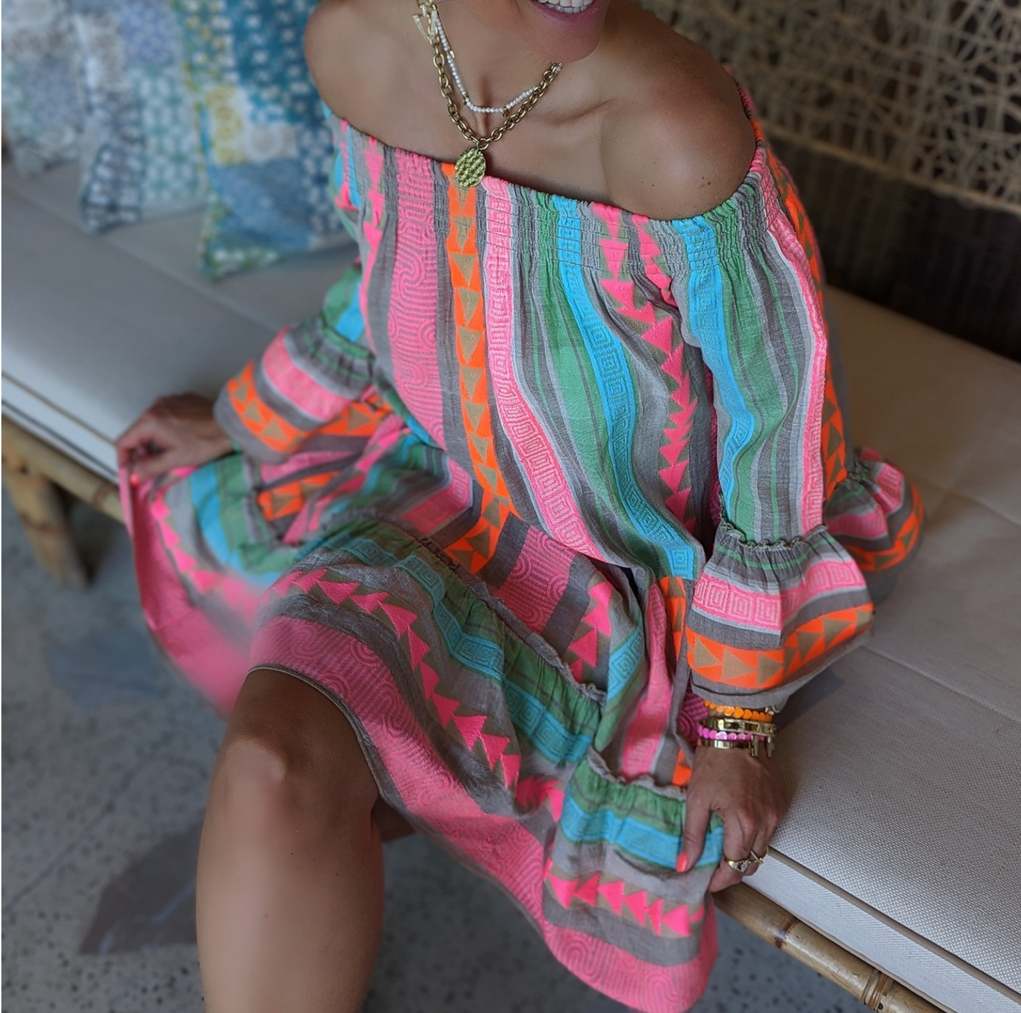 Abito estivo colorato con motivi geometrici, indossato da una donna seduta su un divano in rattan. Stile boho chic, moda femminile estiva.