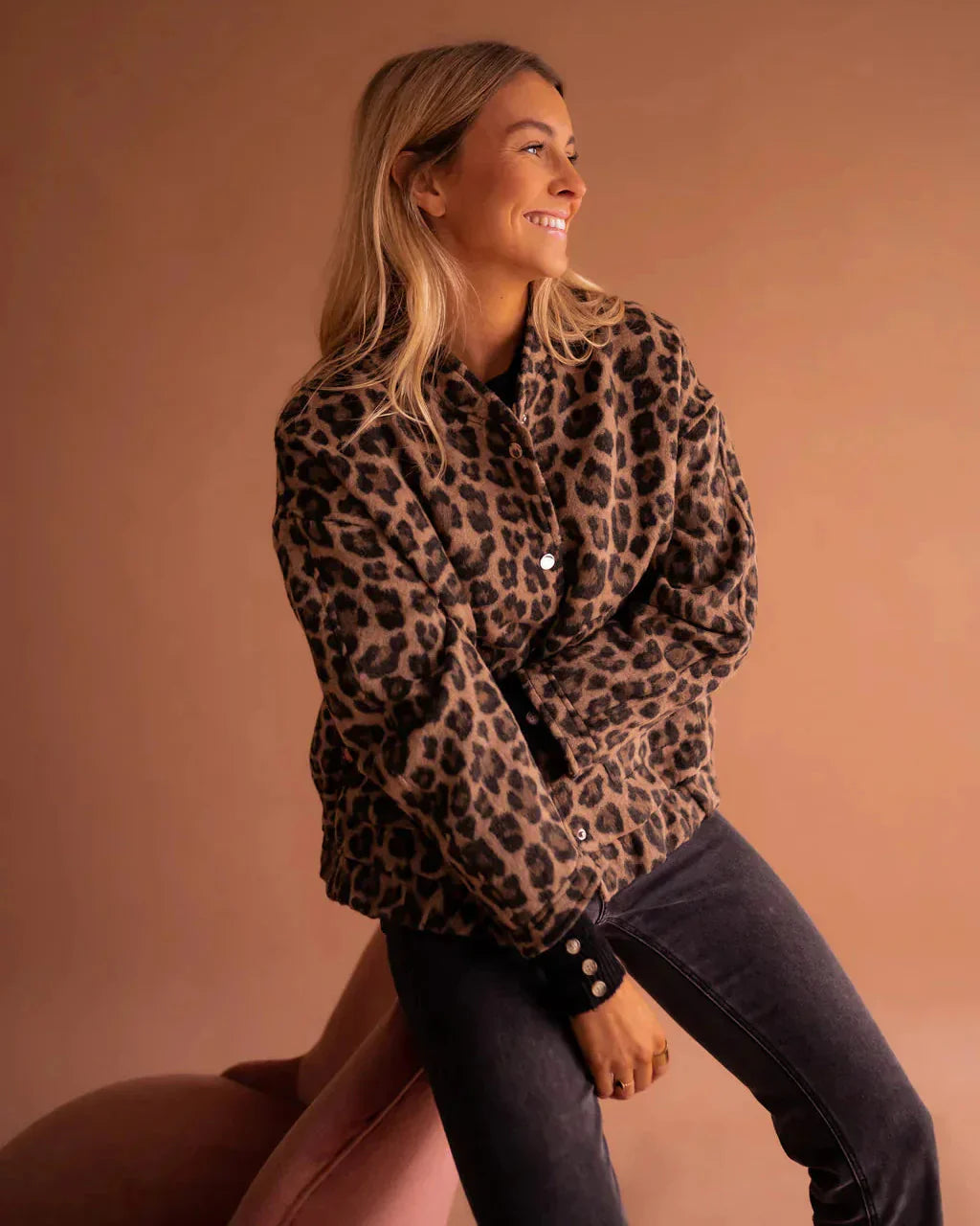 Donna sorridente con giacca leopardata, seduta su sedia rosa, sfondo marrone. Moda autunnale, stile casual chic, tendenze abbigliamento femminile.