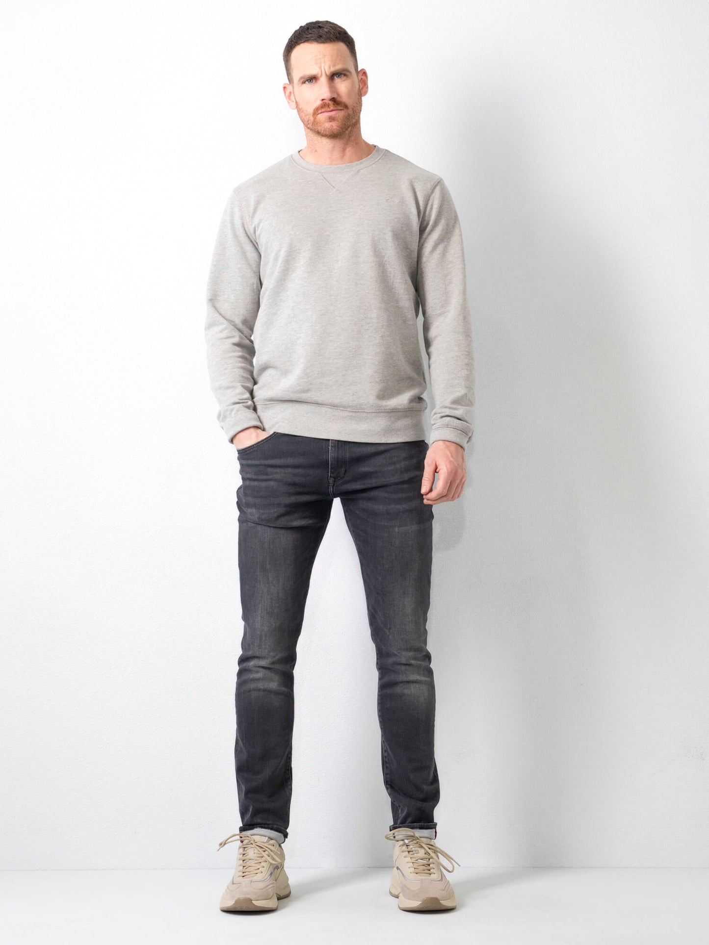 Uomo in posa con maglione grigio e jeans scuri, scarpe beige, sfondo bianco. Moda casual maschile, abbigliamento autunnale, stile moderno.