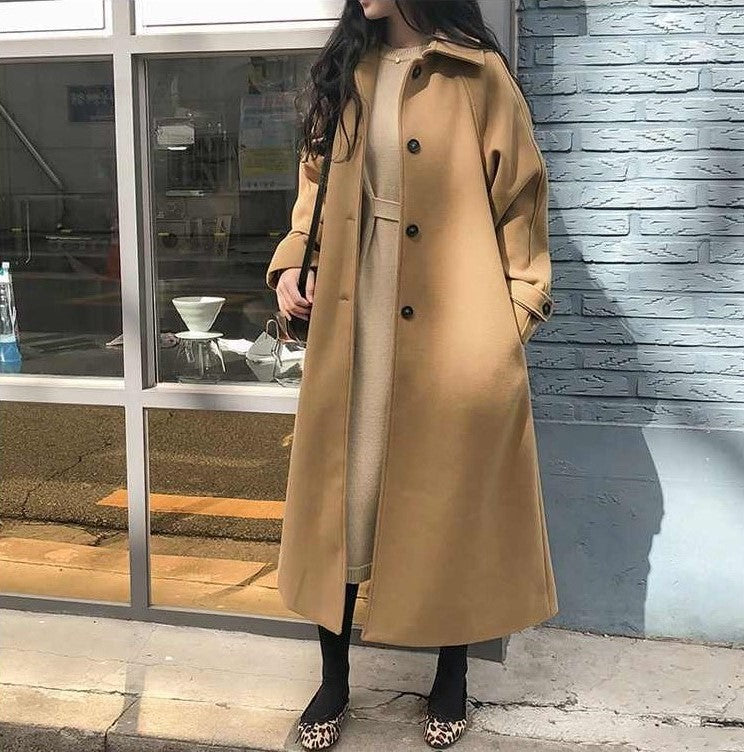 Donna con cappotto lungo color cammello, abito beige, scarpe leopardate, davanti a una vetrina. Moda autunnale, stile elegante e casual.