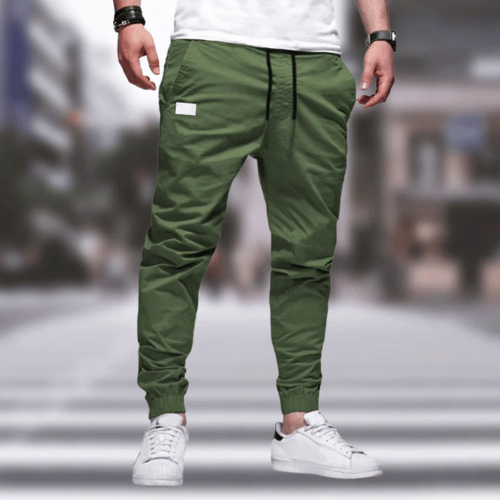 Uomo con pantaloni jogger verdi e scarpe da ginnastica bianche su sfondo urbano sfocato. Moda casual maschile, streetwear moderno.