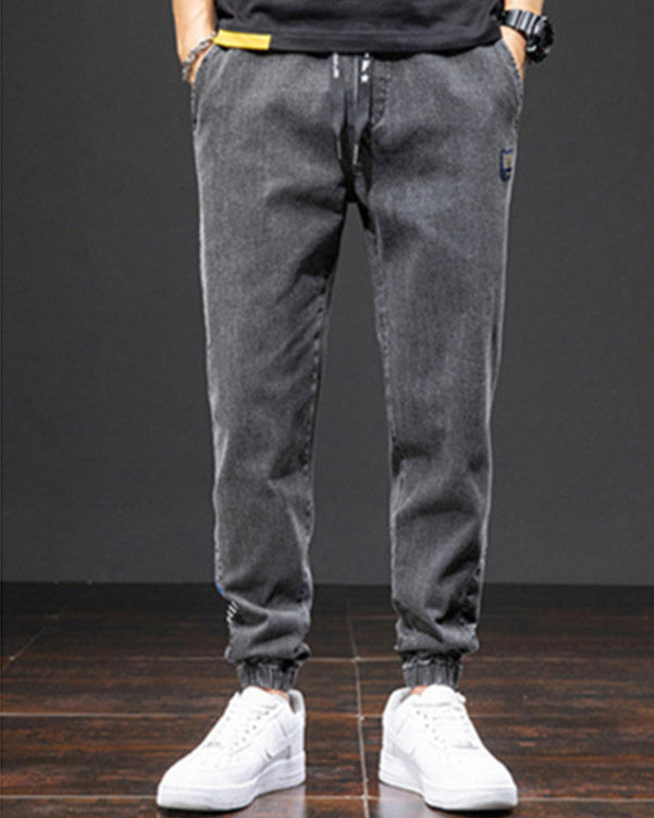 Pantaloni jogger in denim grigio scuro, stile casual, con elastico alla caviglia, indossati con scarpe da ginnastica bianche su sfondo nero.