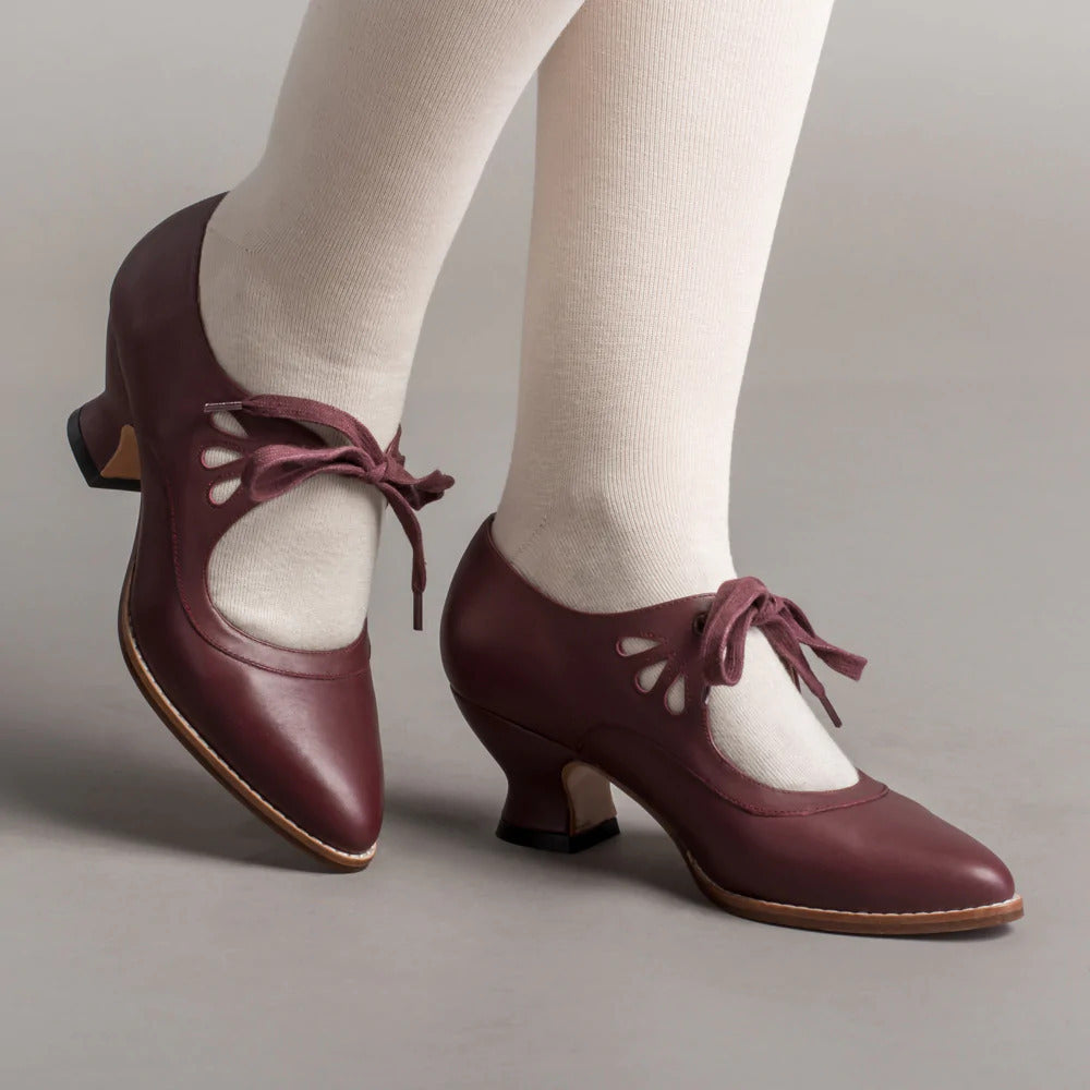 Scarpe vintage in pelle bordeaux con tacco medio e lacci decorativi, indossate con calze bianche. Eleganza retrò, moda anni '20.