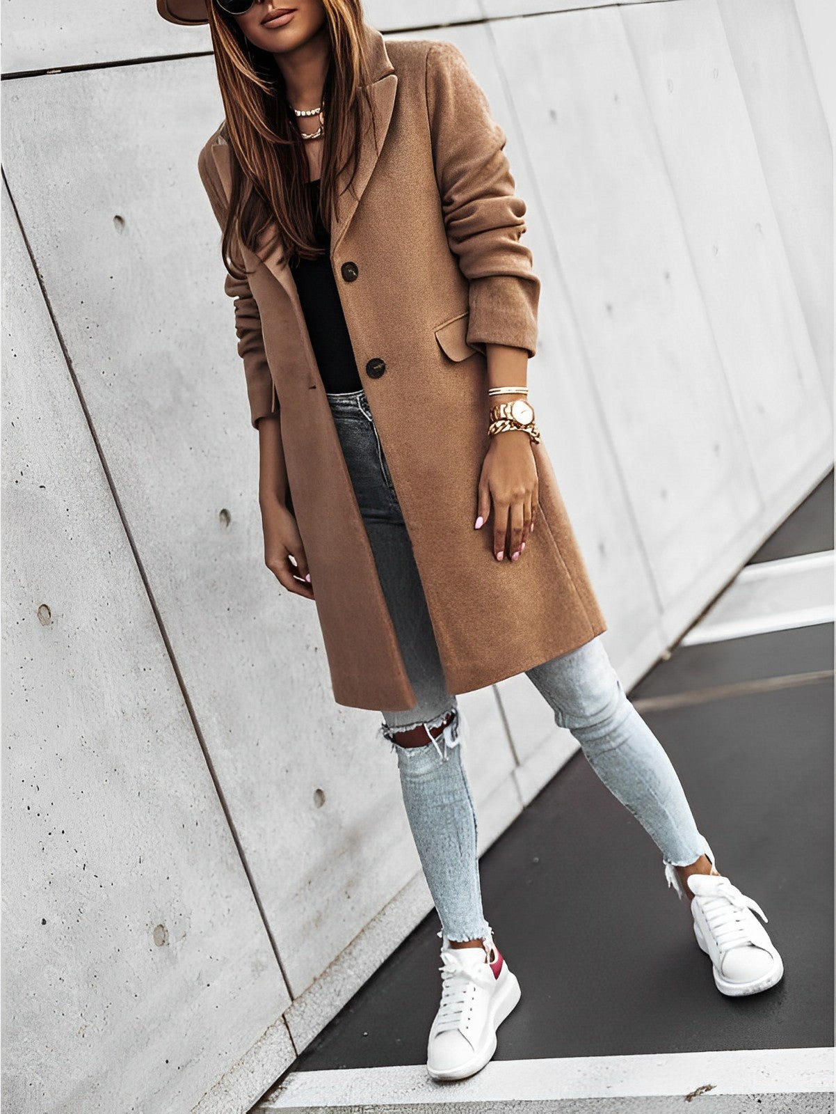 Donna con cappotto marrone, jeans strappati e sneakers bianche, posa contro un muro moderno. Moda autunnale, stile urbano chic.