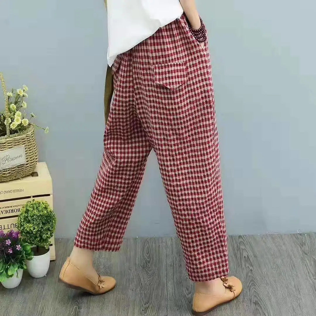 Pantaloni a quadri rossi e bianchi, stile casual, indossati con scarpe marroni su pavimento in legno, sfondo grigio con piante decorative.