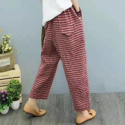 Pantaloni a quadri rossi e bianchi, stile casual, indossati con scarpe marroni su pavimento in legno, sfondo grigio con piante decorative.