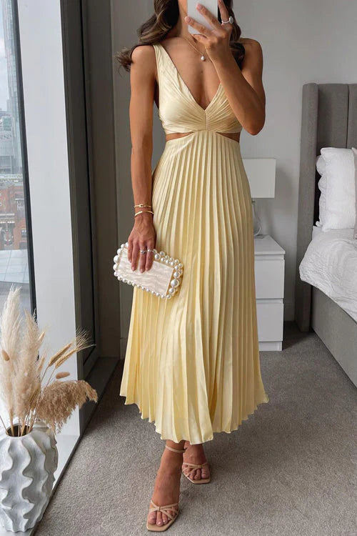 Donna in abito lungo giallo plissettato con scollo a V, sandali beige, borsa bianca con perle, in una camera moderna con finestra panoramica.