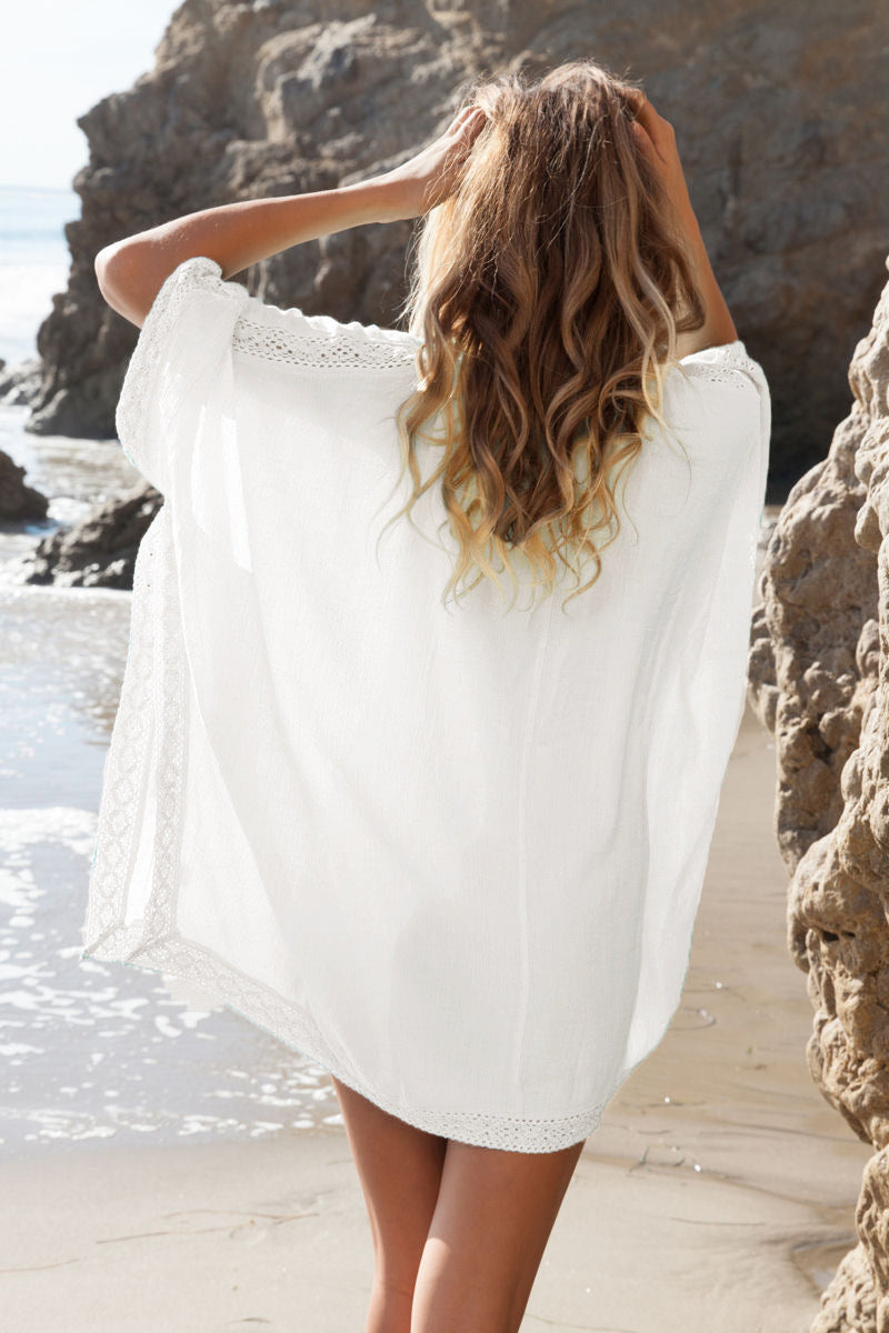 Donna in spiaggia con copricostume bianco, capelli ondulati, scogli sullo sfondo. Moda estiva, relax al mare, abbigliamento leggero.