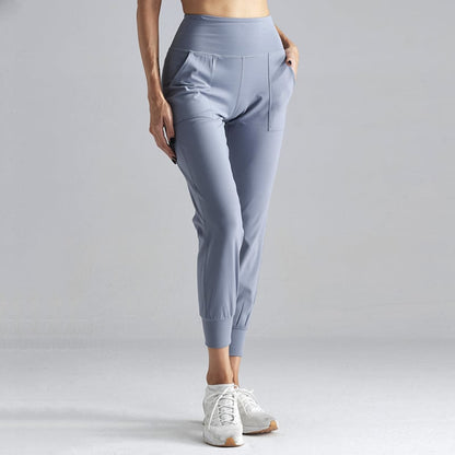 Pantaloni sportivi donna grigi, vita alta, tasche laterali, tessuto elasticizzato, abbinati a scarpe da ginnastica bianche su sfondo neutro.