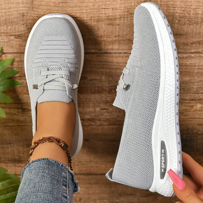 Scarpe da ginnastica grigie da donna su sfondo di legno, design traspirante e suola bianca, indossate con jeans e cavigliera.