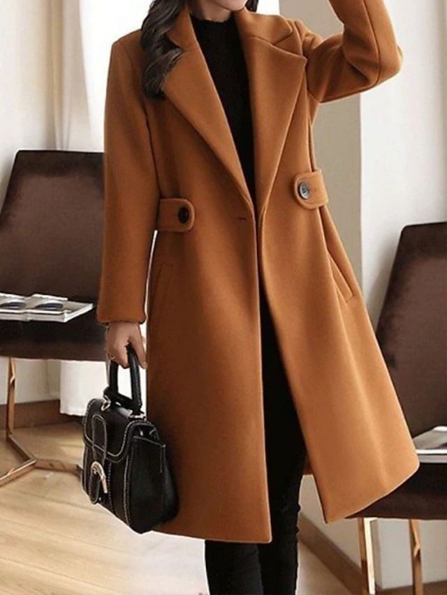 Cappotto lungo color cammello elegante, donna in piedi con borsa nera, moda autunnale, interno moderno, abbigliamento invernale chic.