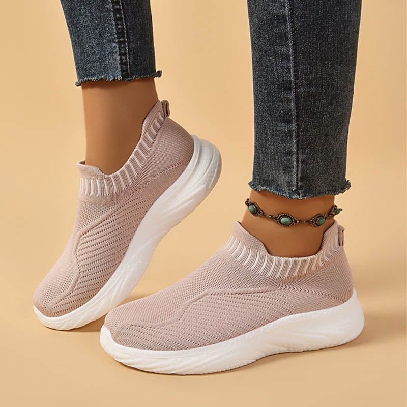 Scarpe da ginnastica rosa chiaro senza lacci, suola bianca, indossate con jeans aderenti e cavigliera decorativa. Moda casual donna.