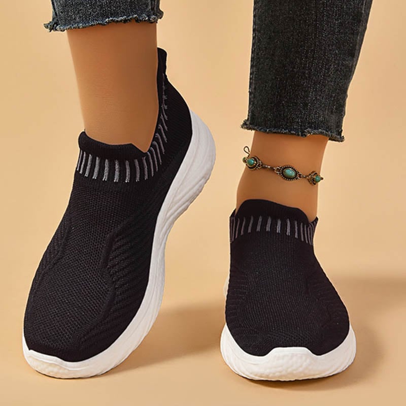 Scarpe da ginnastica nere senza lacci con suola bianca, indossate con jeans e cavigliera verde. Scarpe sportive comode e alla moda.