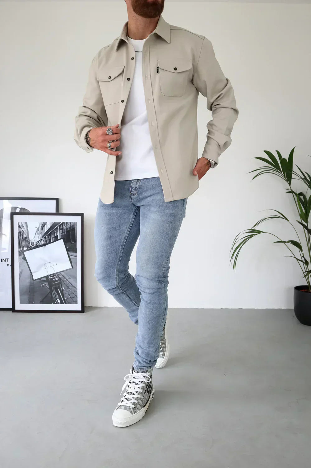 Uomo in camicia beige e jeans chiari, indossa sneakers bianche con motivo. Sfondo minimalista con piante e quadri. Moda casual maschile.