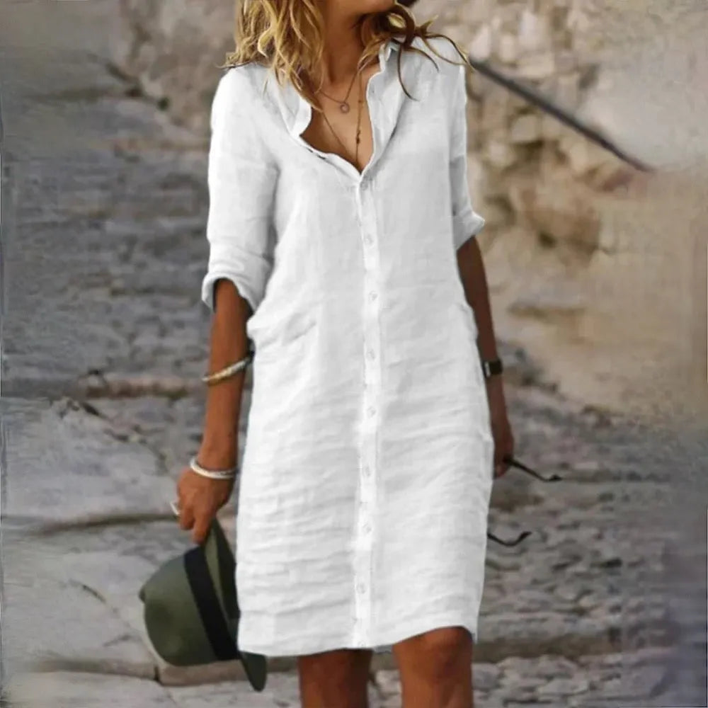 Donna con abito bianco di lino, maniche lunghe, cammina su strada acciottolata. Moda estiva casual, stile elegante, accessori minimal.