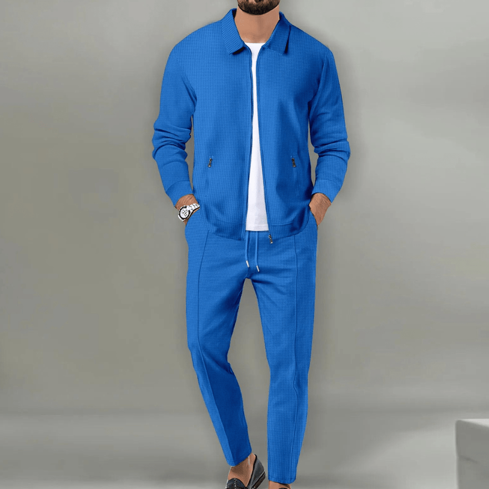 Uomo in abito sportivo blu, giacca e pantaloni coordinati, sfondo grigio. Moda maschile elegante, stile casual-chic, abbigliamento moderno.