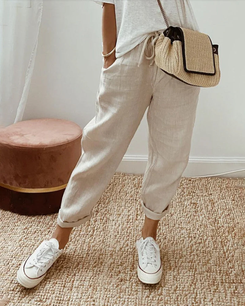 Pantaloni di lino beige, scarpe da ginnastica bianche, borsa a tracolla in paglia, moda casual estiva, donna, pavimento in moquette, sgabello rosa.