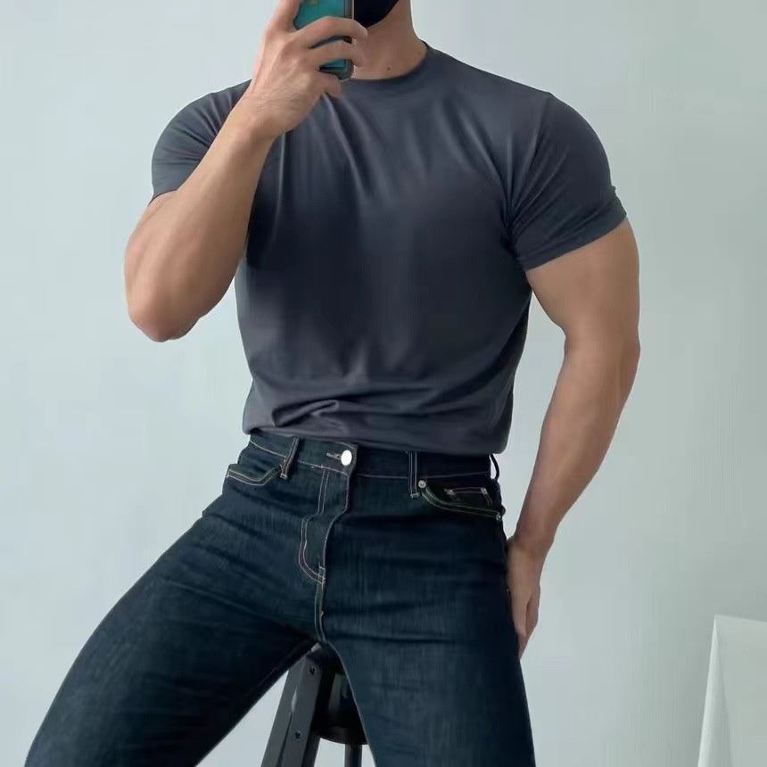 Uomo muscoloso in maglietta grigia e jeans scuri, seduto su sgabello, scatta selfie con smartphone. Moda casual maschile, fitness, abbigliamento moderno.