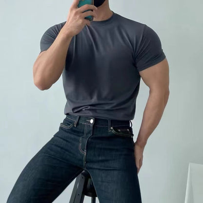 Uomo muscoloso in maglietta grigia e jeans scuri, seduto su sgabello, scatta selfie con smartphone. Moda casual maschile, fitness, abbigliamento moderno.