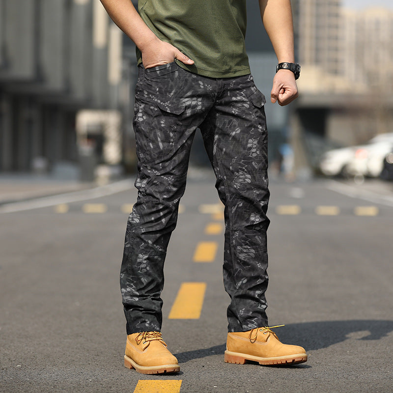 Uomo in piedi su strada indossando pantaloni mimetici neri e scarpe gialle, sfondo urbano sfocato. Moda maschile casual, stile urbano.