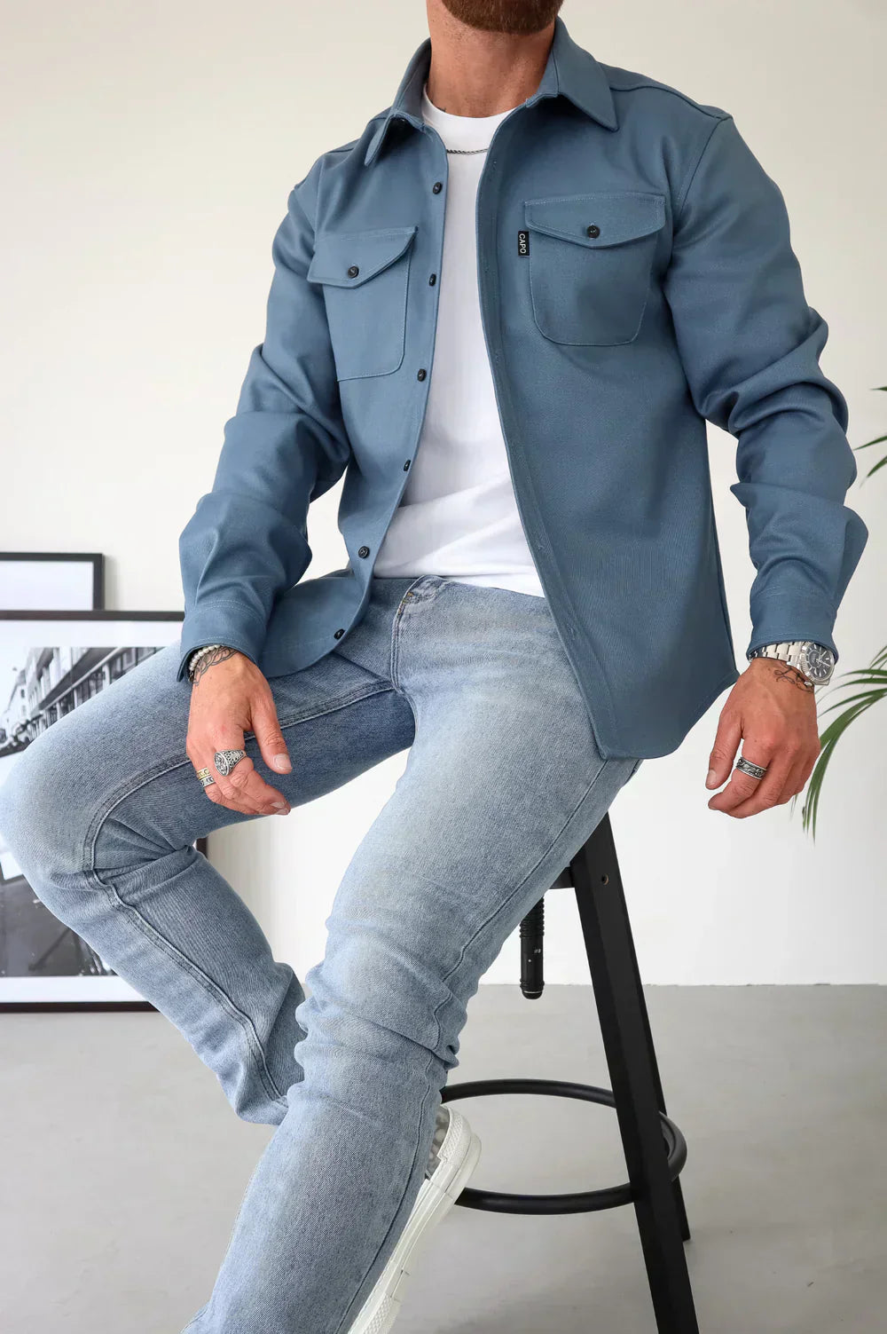 Uomo seduto su sgabello indossa camicia blu con tasche, jeans chiari e orologio argento. Stile casual moderno, moda maschile autunnale.