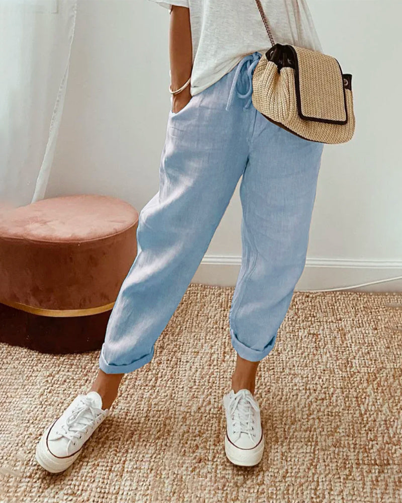 Donna con pantaloni di lino azzurri, borsa a tracolla in paglia e sneakers bianche su tappeto beige. Moda casual estiva, stile rilassato.