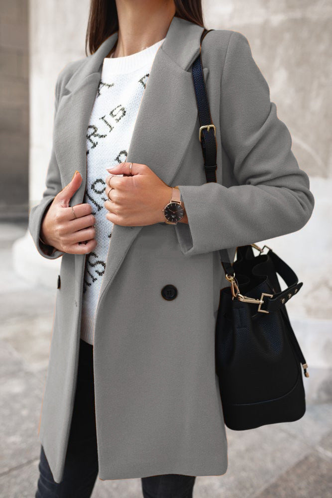 Donna con cappotto grigio elegante, maglione bianco con numeri, orologio da polso nero e borsa a tracolla nera. Moda autunnale chic.