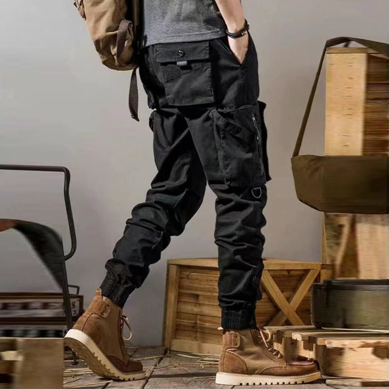 Pantaloni cargo neri da uomo con tasche multiple, abbinati a scarponcini marroni e zaino. Stile casual outdoor, moda maschile autunnale.