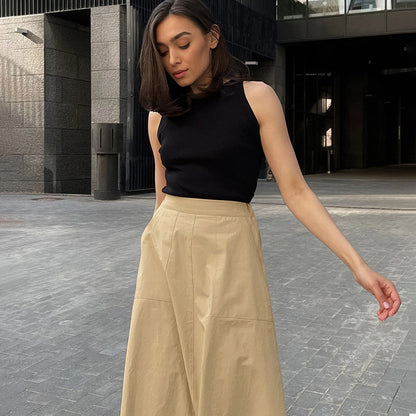 Donna con top nero e gonna beige in posa all'aperto, architettura moderna sullo sfondo. Moda urbana, abbigliamento casual chic, stile contemporaneo.