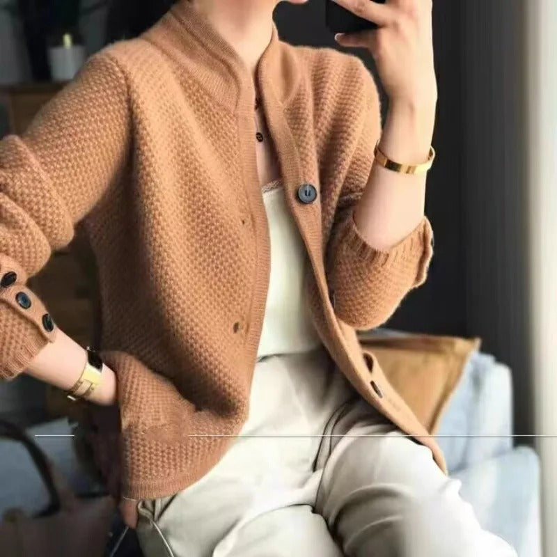 Donna con cardigan marrone a maglia, camicia bianca e pantaloni chiari, seduta vicino a una finestra. Moda autunnale, abbigliamento casual elegante.