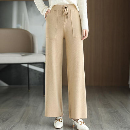 Pantaloni beige a gamba larga in maglia, con tasche frontali e cintura, indossati con maglione bianco e mocassini bianchi su pavimento in legno.