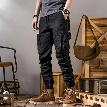 Pantaloni cargo neri da uomo con tasche multiple, abbinati a stivali marroni su pavimento in legno. Moda casual maschile, stile urbano.