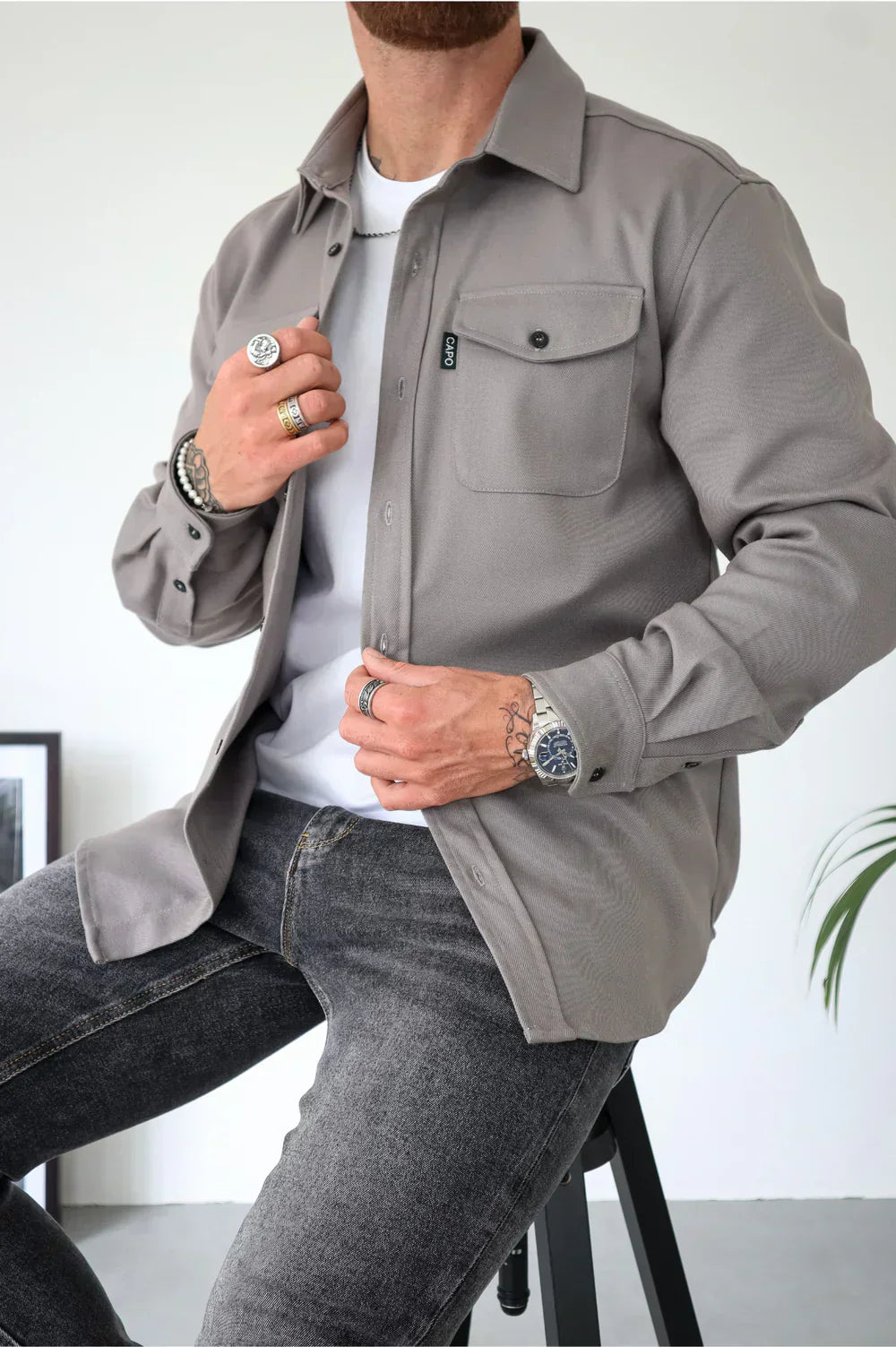 Uomo con camicia grigia casual, jeans scuri, orologio e anelli, seduto su sgabello. Moda maschile, abbigliamento moderno, accessori eleganti.