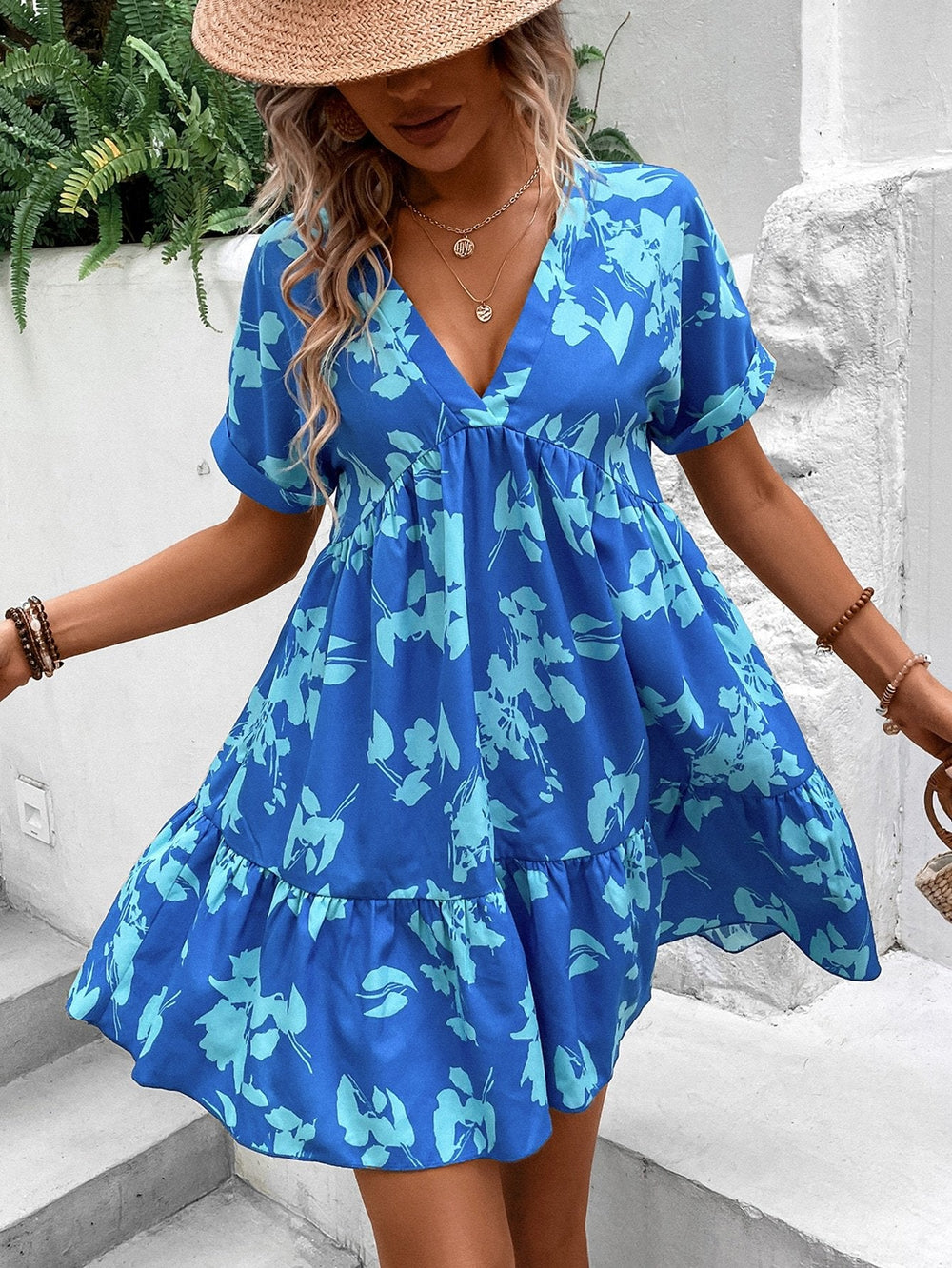 Donna con abito estivo blu a fiori, cappello di paglia, collana e bracciali, su sfondo di piante verdi. Moda estiva, abbigliamento femminile elegante.