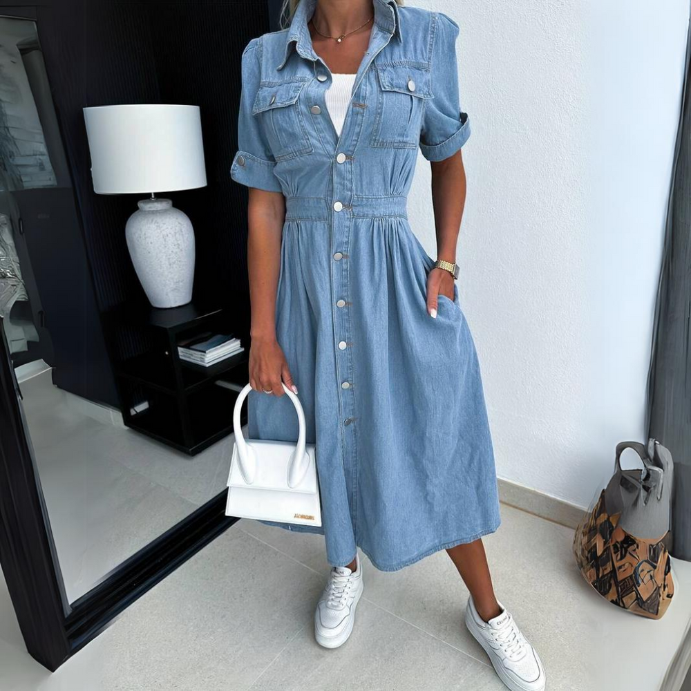 Donna in abito di jeans con bottoni, borsa bianca e sneakers bianche, accanto a specchio e lampada moderna. Moda casual chic.