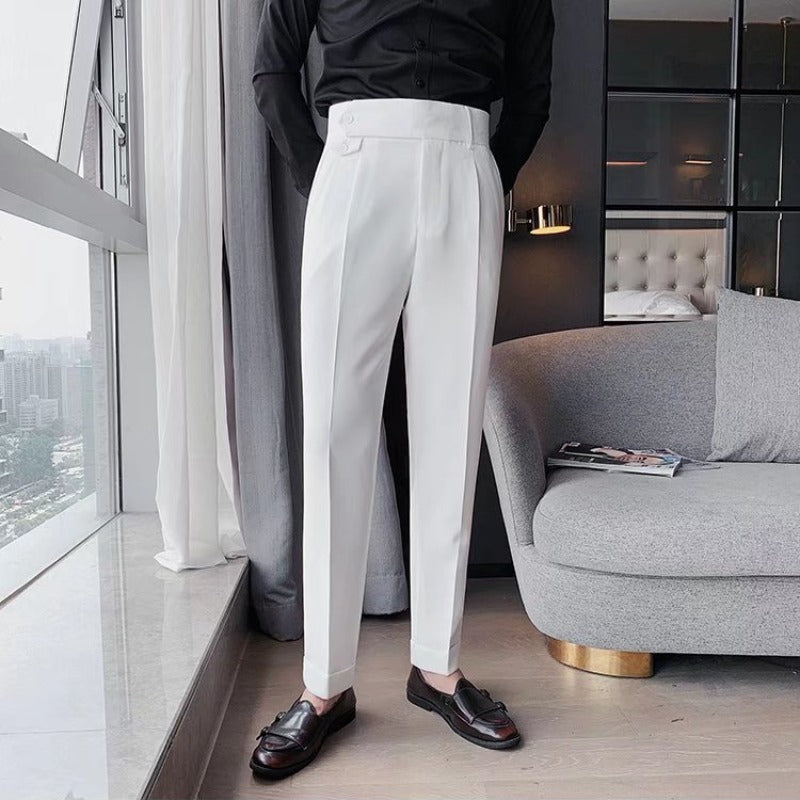 Uomo in pantaloni bianchi eleganti, camicia nera, mocassini marroni, in un salotto moderno con divano grigio e finestra panoramica.