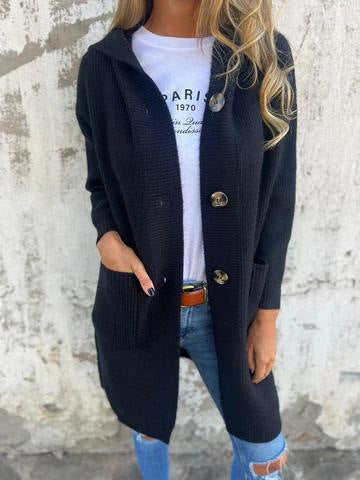 Donna con cardigan nero lungo, bottoni grandi, tasche frontali, indossa jeans strappati e maglietta bianca con scritta, sfondo muro grigio.