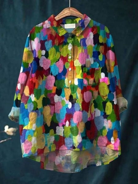 Camicia colorata a maniche lunghe con motivo astratto multicolore su sfondo blu scuro, appesa a una gruccia di legno. Moda vivace e artistica.