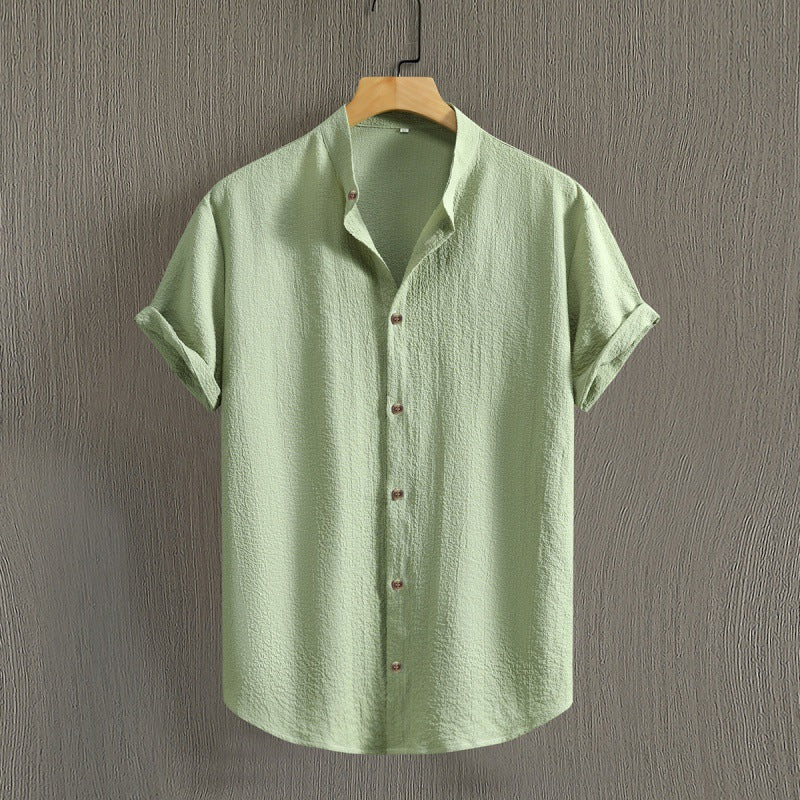 Camicia verde chiaro in lino, maniche corte, colletto classico, bottoni frontali, appesa su gruccia di legno, sfondo grigio testurizzato.