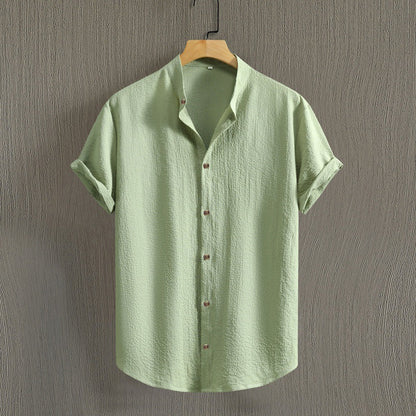 Camicia verde chiaro in lino, maniche corte, colletto classico, bottoni frontali, appesa su gruccia di legno, sfondo grigio testurizzato.