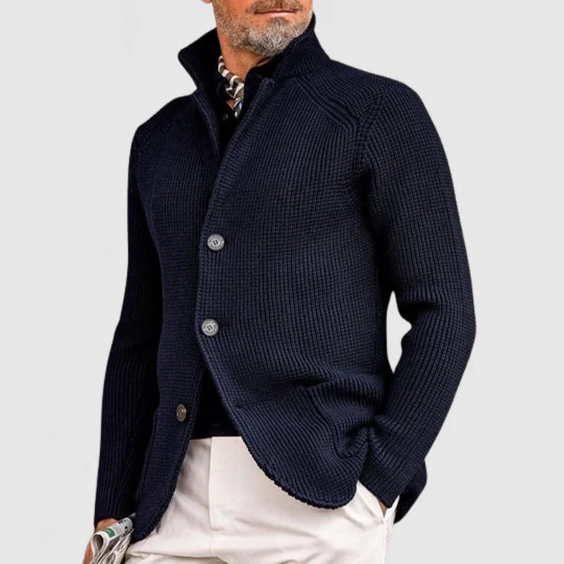 Uomo con maglione blu scuro a maglia grossa, colletto alto, abbinato a pantaloni chiari. Moda maschile elegante, stile autunnale.