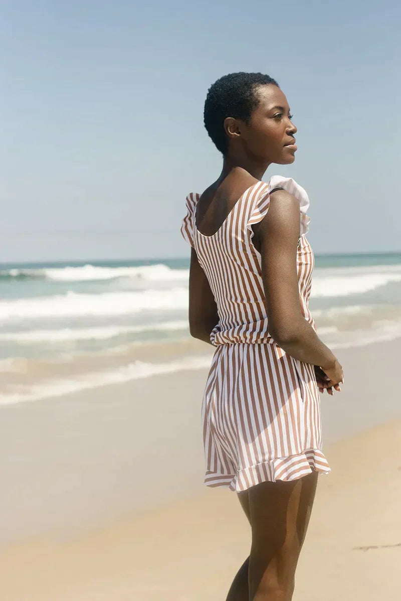 Donna in spiaggia con abito a righe bianche e marroni, mare sullo sfondo. Moda estiva, vacanza al mare, abbigliamento casual elegante.