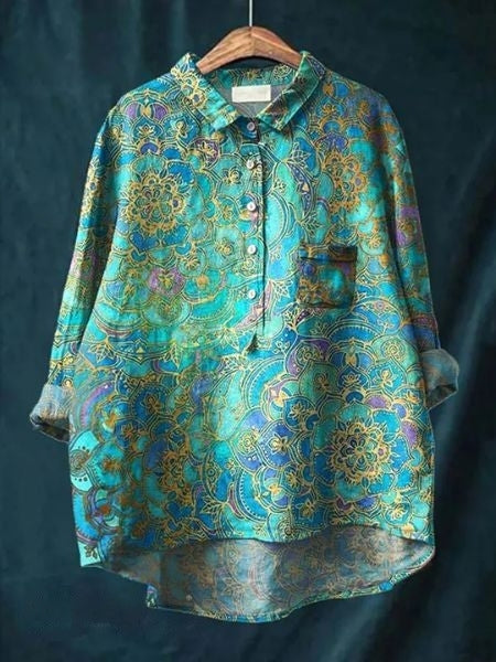 Camicia oversize in seta blu con motivi floreali dorati, maniche lunghe, colletto classico e taschino, appesa su sfondo scuro.