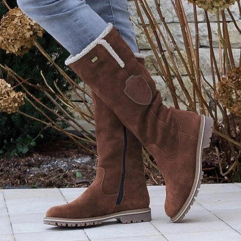 Stivali invernali alti marroni in pelle scamosciata con fodera in pelliccia, indossati con jeans, su sfondo di piante secche e muro di pietra.
