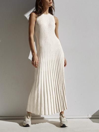 Donna in abito lungo plissettato bianco, senza maniche, con sneakers bianche, posa contro un muro bianco. Moda estiva elegante e casual.