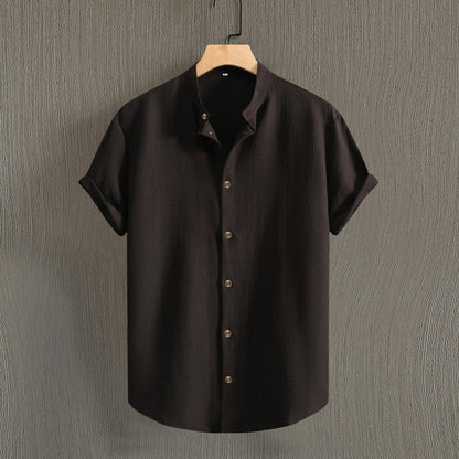 Camicia nera a maniche corte in cotone, design minimalista, bottoni frontali, appesa su gruccia di legno, sfondo grigio testurizzato.