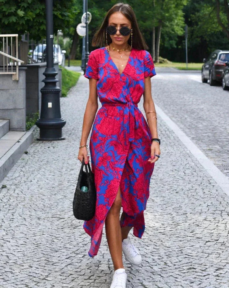 Donna con abito floreale rosso e blu, occhiali da sole, borsa nera, cammina su strada acciottolata. Moda estiva, look casual chic.