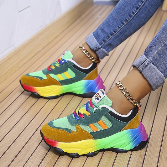 Scarpe da ginnastica multicolore con suola arcobaleno, lacci colorati e dettagli verdi, indossate con jeans e cavigliere dorate su pavimento in legno.